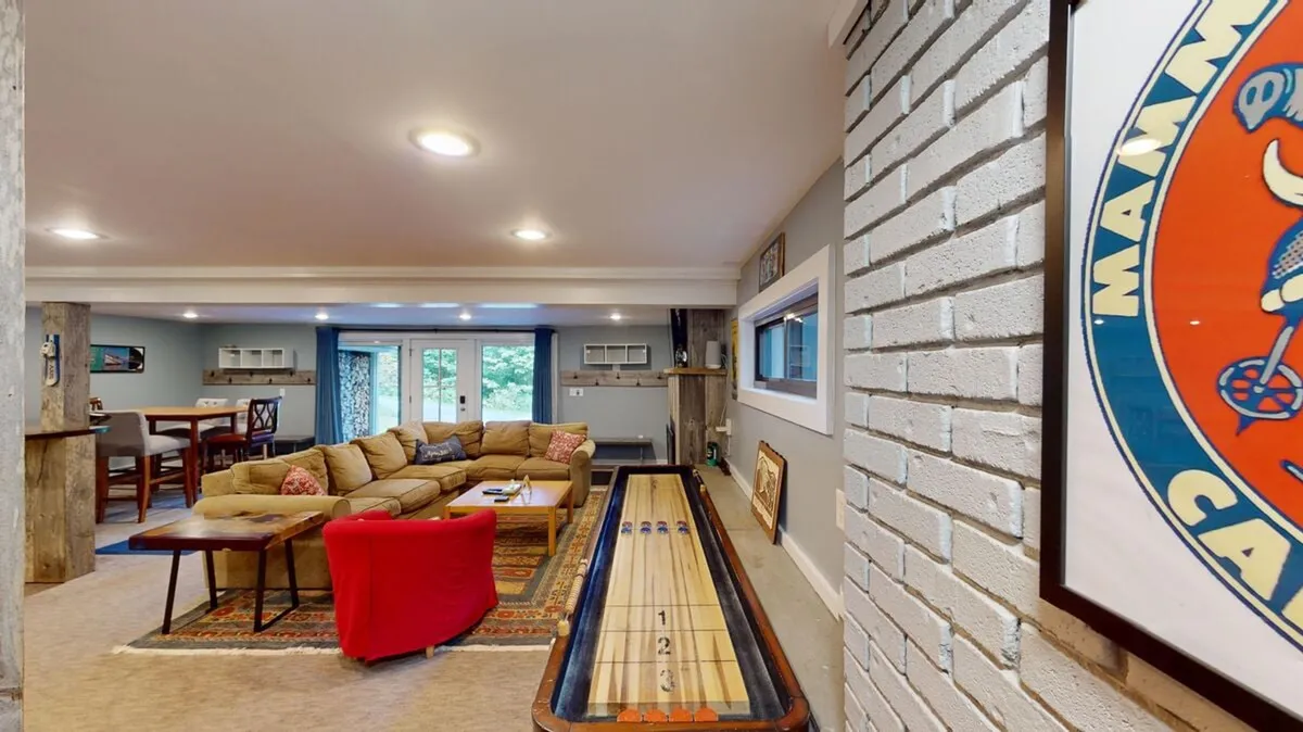 A light-filled recreation area featuring a shuffleboard table, a colorful rug, comfortable sofa, and large windows bringing in natural light. The setup is modern and designed for both play and relaxation, with a clear emphasis on family-friendly fun.