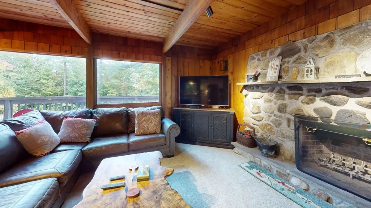 A cozy living room with a stone fireplace, large leather sofa, wooden coffee table, television, and expansive windows looking out onto nature. The warm, inviting space is perfect for unwinding and socializing year-round.