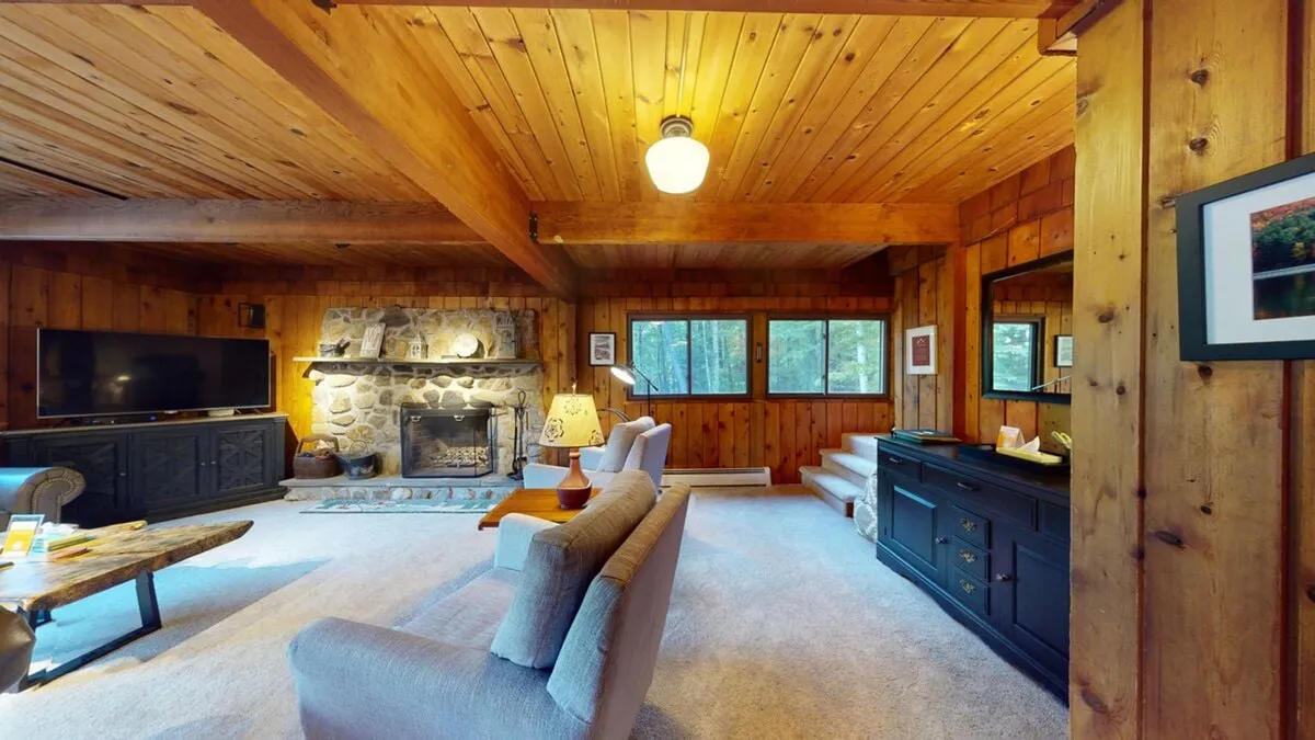 A welcoming living/media area featuring a stone fireplace, wooden beams, comfortable seating, a large television, and expansive windows. The room is arranged for group enjoyment, movie nights, and socializing after a day outdoors.