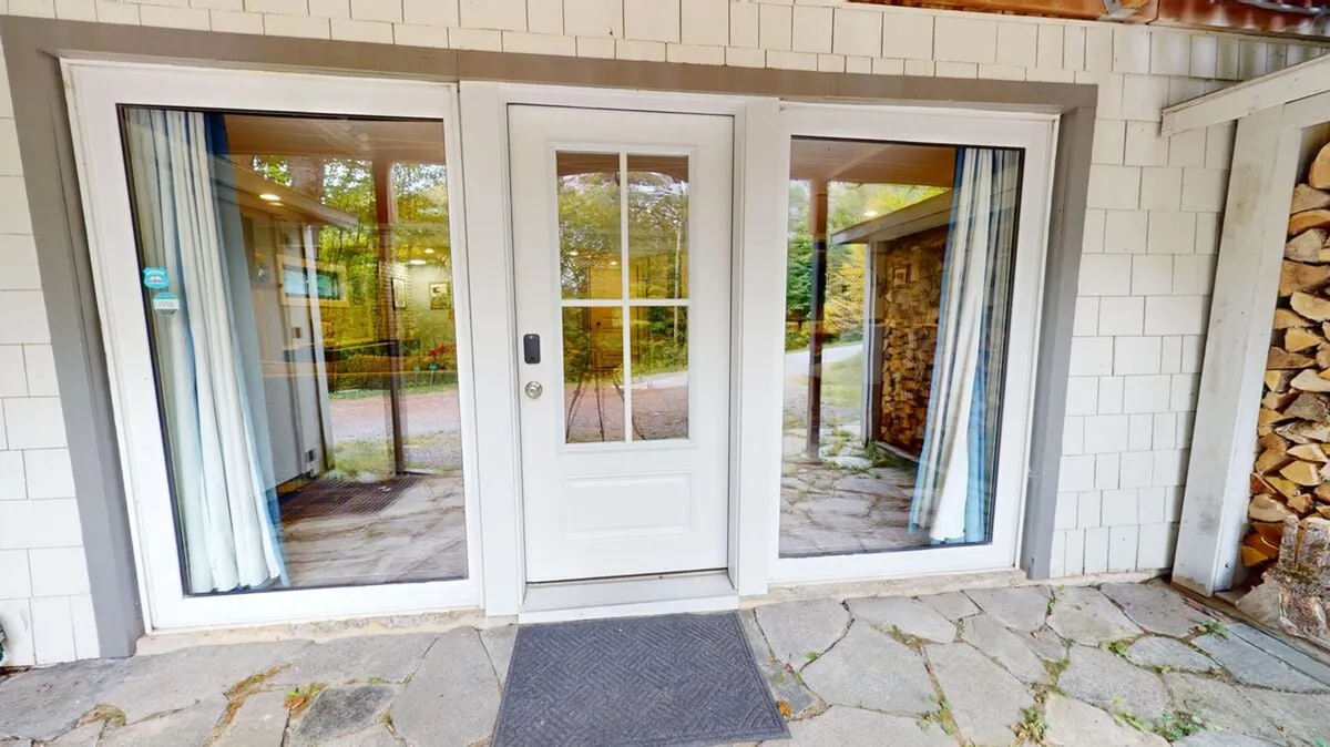 The image features a charming front entrance with large glass windows, a stone pathway, and a neatly stacked woodpile, nestled among trees. This view emphasizes the harmonious integration of the home with its natural surroundings and hints at the stonework mentioned in the section. The visual transition from the outside in underscores year-round enjoyment and a seamless connection to nature.