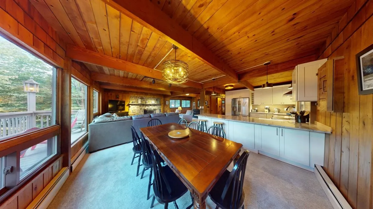 A wide perspective of the main living area displays a dining table, open kitchen, and cozy living space with ample windows. The interconnectedness of the spaces is apparent, with the dining area and seating positioned for easy conversation and group activities.