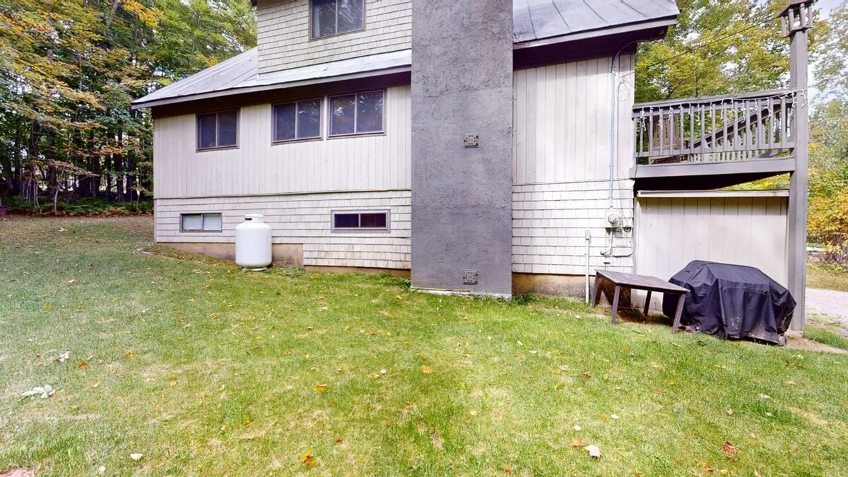 A calm backyard scene featuring green grass, a deck area, and an inviting exterior wall of the home. The deck provides a space for morning coffee or evening relaxation, while the yard offers a private area for outdoor gatherings, bordered by trees for privacy.