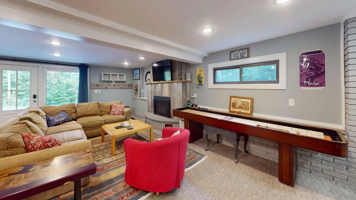 The image showcases a welcoming living room anchored by a fireplace, with a comfortable sofa, colorful cushions, a coffee table, and a game table. Artwork on the walls and casual seating invite group relaxation, storytelling, and leisurely evenings.