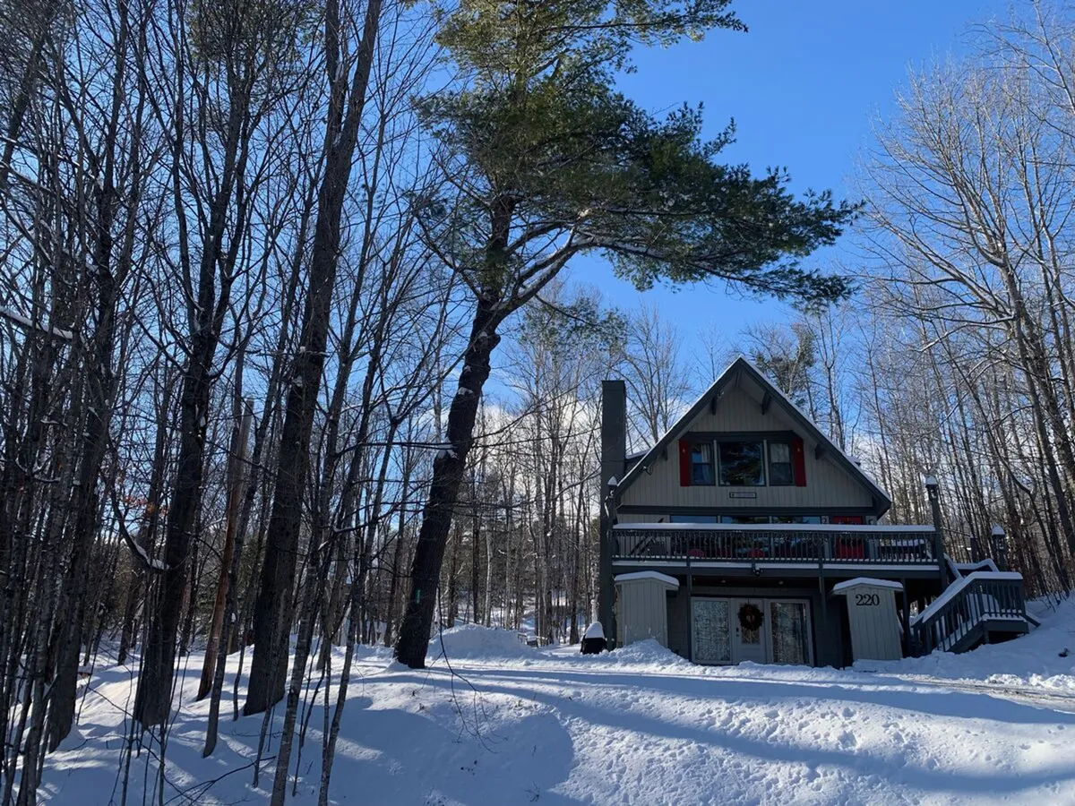 An A-frame cabin stands in a snowy landscape surrounded by tall trees, suggesting both winter activities and the comfort of a mountain getaway. The inviting porch and snow-covered ground evoke post-adventure relaxation and group gatherings.