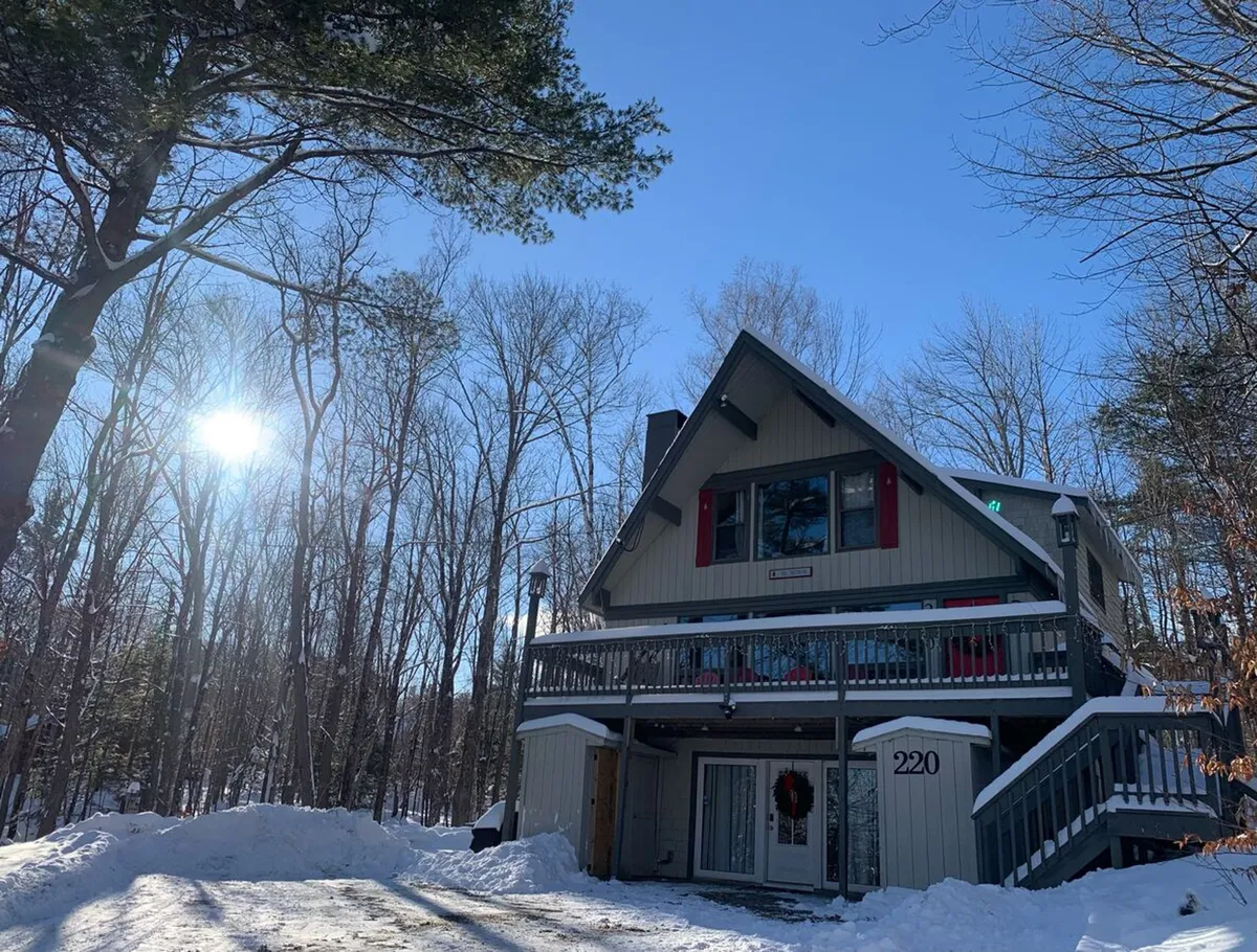 This image captures the exterior of the property in winter, showing the house nestled among trees, with the ground blanketed in snow and the sun shining brightly. The scene evokes a sense of immediate access to snowy adventure and a cozy mountain retreat.