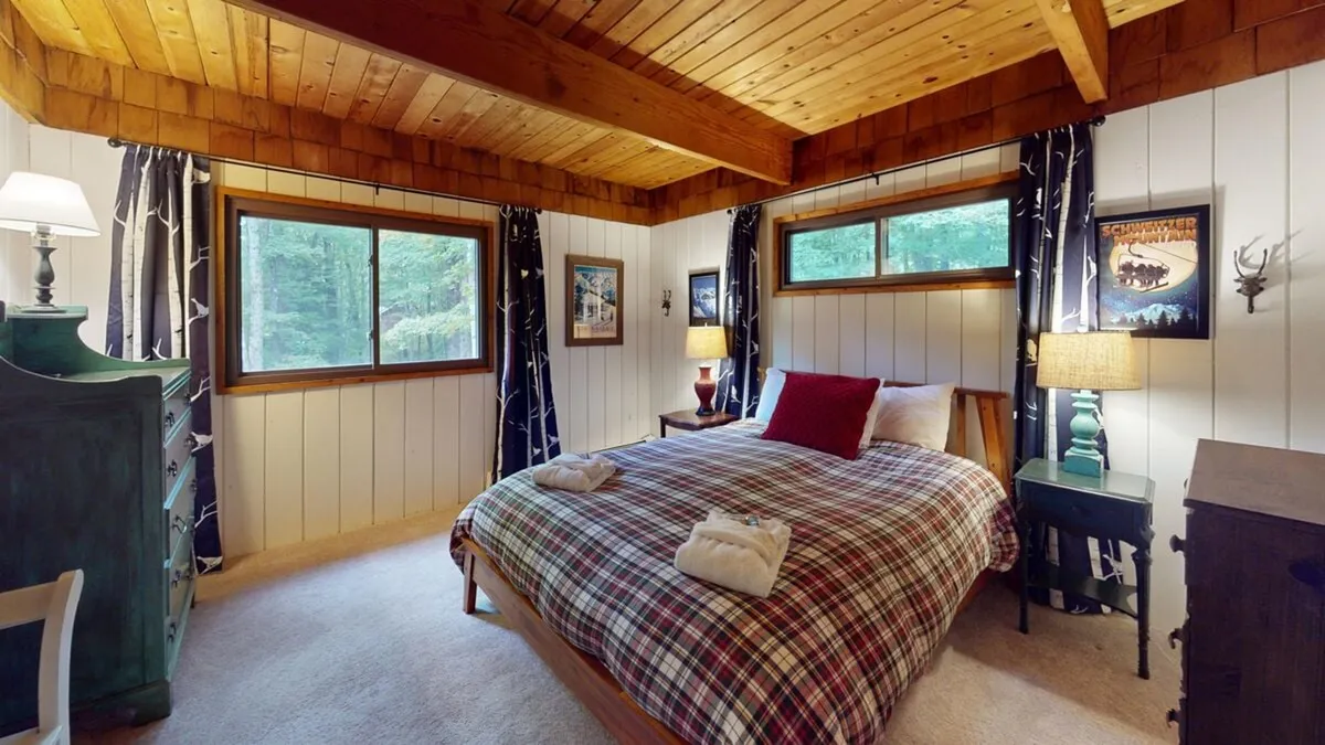 A serene bedroom with a queen or full bed adorned in a plaid bedspread, wooden beams overhead, and windows offering calming forest views. Soft decorative lamps and tasteful wall art contribute to the restful atmosphere.