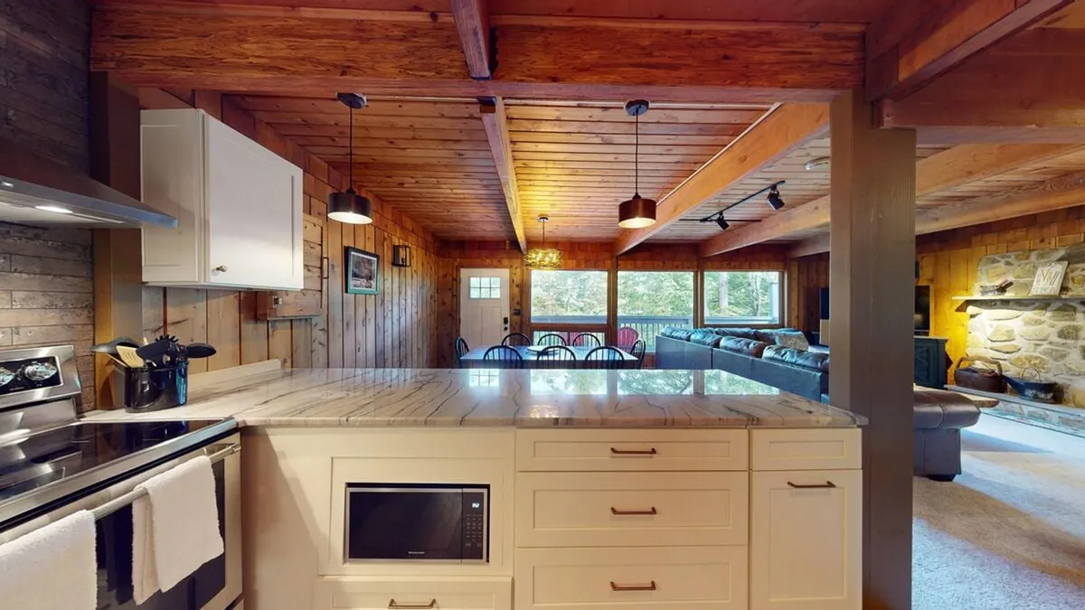 This image features a spacious and beautifully appointed kitchen, complete with a large island, stainless steel appliances, wooden beams, and large windows that let in ample natural light. The stone fireplace in the background connects the kitchen to the cozy living area, hinting at a seamless open-concept layout ideal for both cooking and socializing.