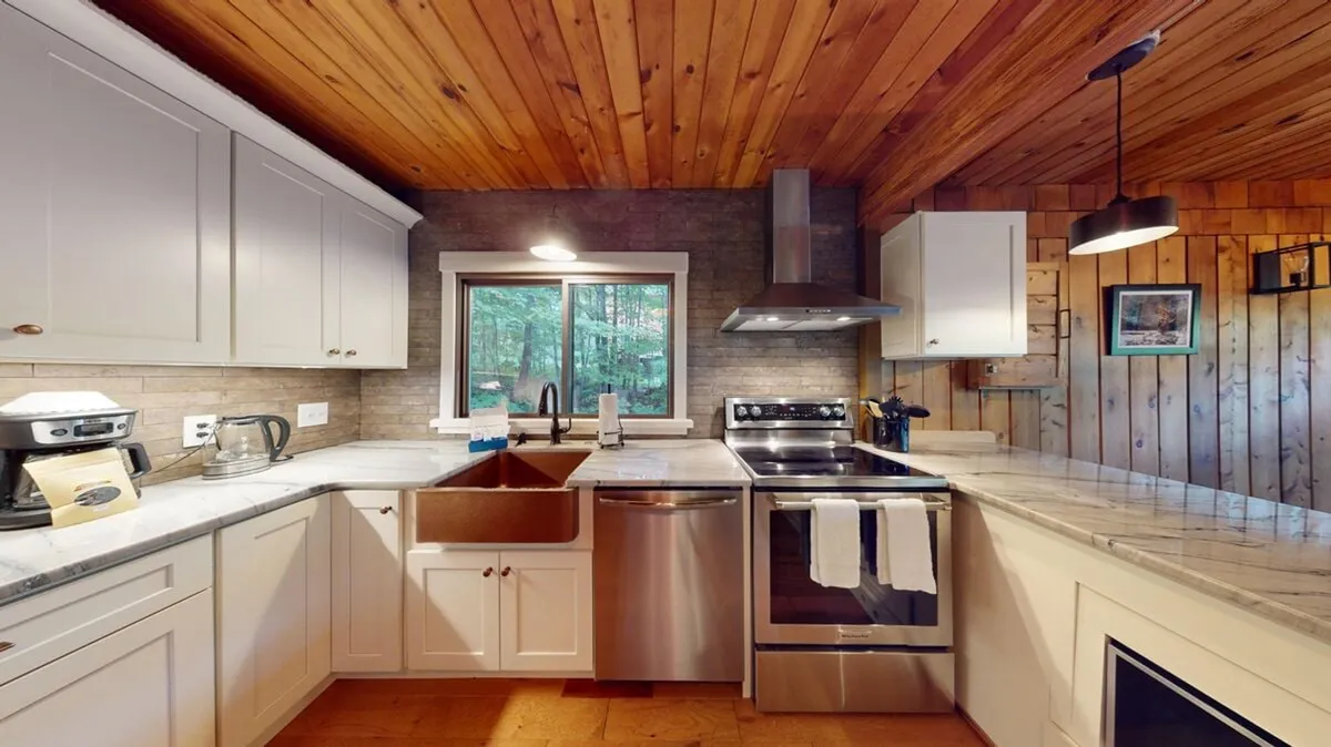 The image showcases a bright, inviting kitchen with a marble countertop, sleek stainless steel appliances, and a large window that frames the tranquil forest views outside. The wooden ceiling adds a cozy, chalet-style warmth, while the modern sink and open layout suggest a space perfect for gatherings and shared meals.