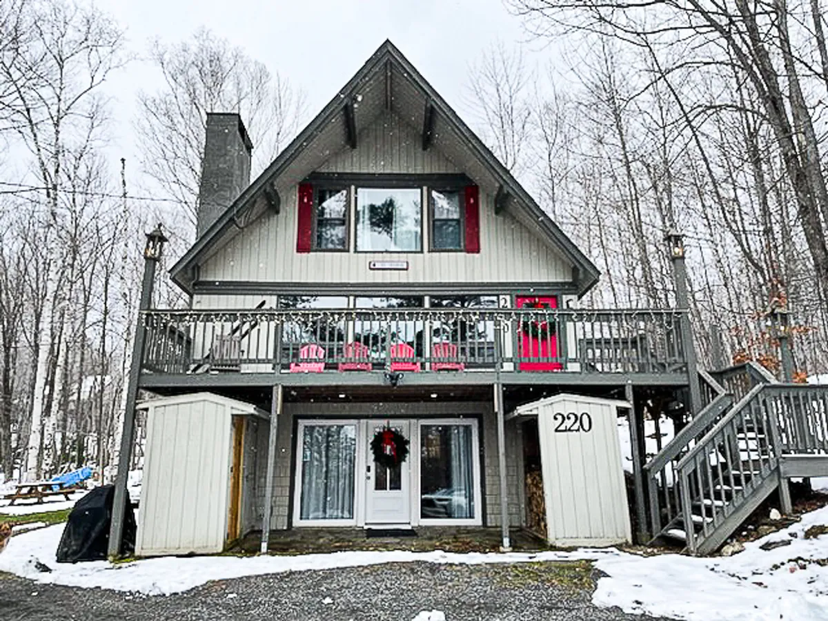 A charming A-frame chalet stands amidst snowy trees, its front balcony adorned with bright red chairs and a festive wreath on the door. The inviting exterior showcases the chalet’s unique architecture and hints at the coziness within, all set in a tranquil winter landscape.