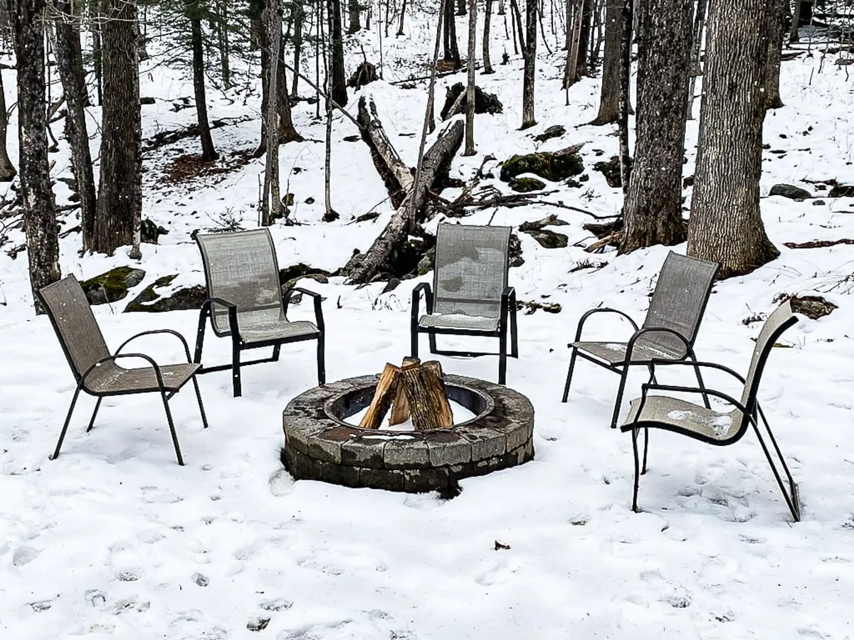 An inviting outdoor fire pit encircled by multiple chairs sits amidst gently falling snow and tall trees, creating a warm, communal space for group gatherings and storytelling. The surrounding wooded area offers privacy and a sense of escape, perfectly in line with the chalet's serene setting.